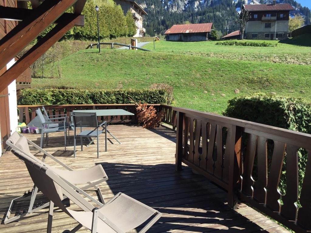 A wooden terrace is depicted with several outdoor furniture items, including a dining table and chairs, as well as lounge chairs. The scene overlooks a green landscape with distant buildings and mountains in the background, creating a tranquil outdoor setting.