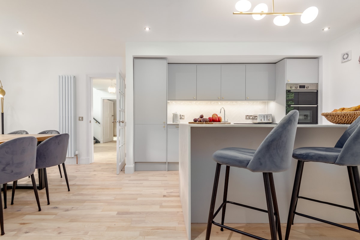 A bright and modern kitchen space is equipped with sleek cabinetry and a spacious island. Bar stools provide seating for casual dining. The counter is adorned with fresh fruit, and the dining area features a wooden table with upholstered chairs.