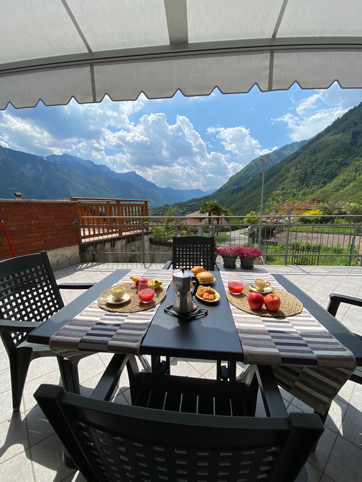 A spacious terrace is presented, featuring a dining table set with cups, a coffee pot, fruit, and pastries. The backdrop showcases dramatic mountain views under a partially cloudy sky. Colorful flowers line the terrace, enhancing the serene outdoor setting.