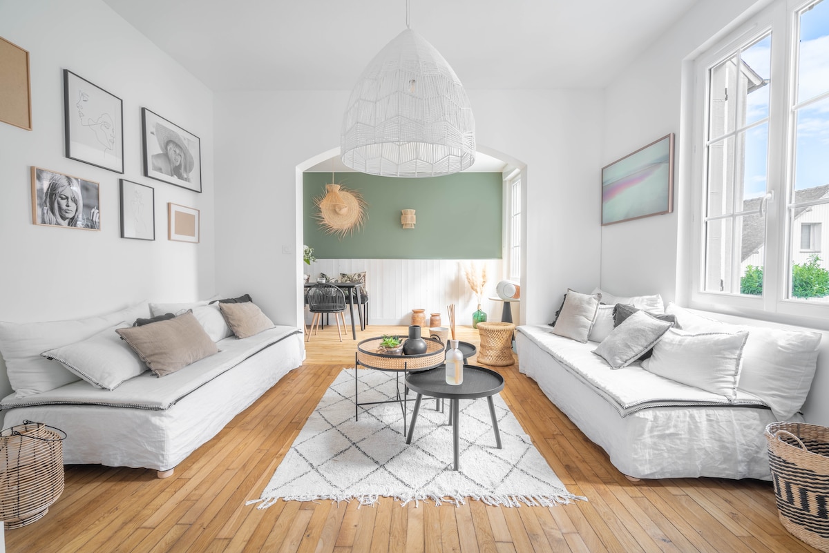 The living area features two light-colored sofas positioned around a low round table. Natural light enters through large windows, highlighting wooden flooring. Decorative elements include framed artwork on the walls and textured woven baskets, contributing to a contemporary yet comfortable environment.