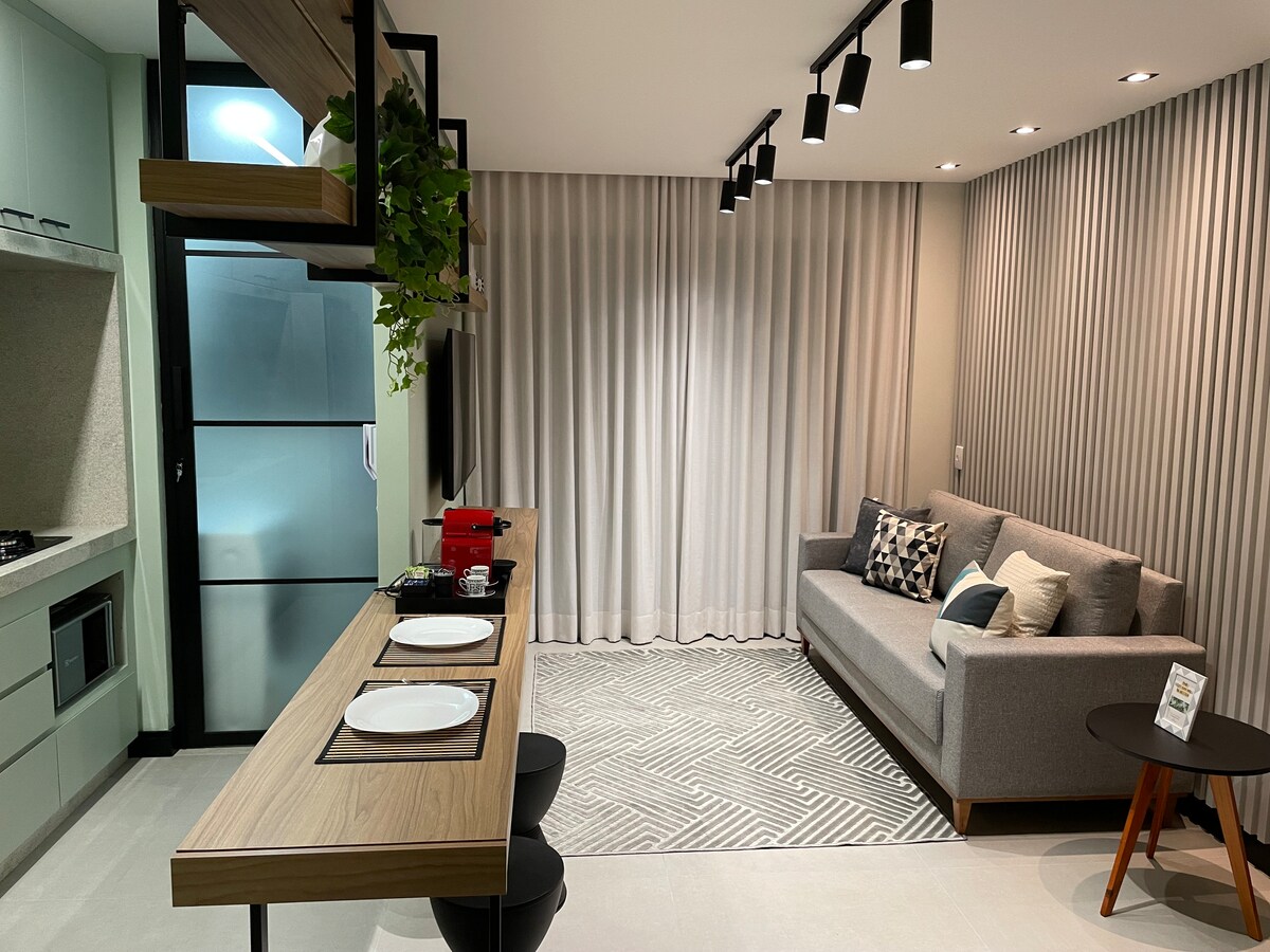 A modern living space features a cozy gray sofa paired with decorative cushions. A dining table with place settings sits opposite an open kitchen showcasing sleek cabinetry. Soft curtains filter natural light, while a tasteful area rug adds texture to the floor.