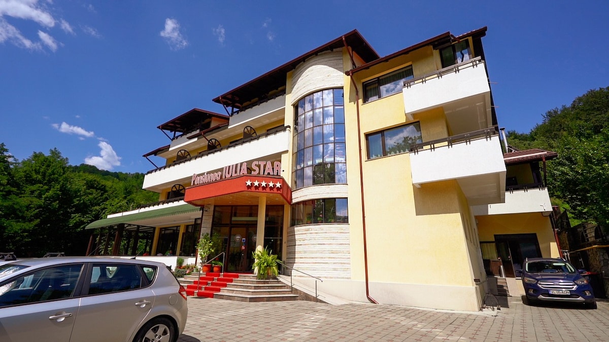 The three-story building features a prominent entrance with large glass windows and a welcoming facade. The surrounding area includes well-maintained greenery and a parking lot visible in the foreground. Bright sunlight enhances the colors of the building against a clear blue sky.