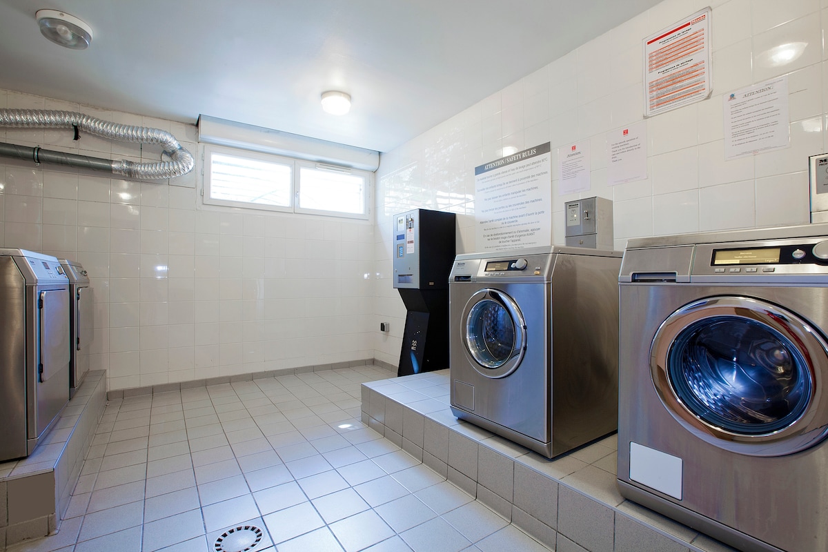 A clean laundry room is equipped with multiple stainless steel washing machines and dryers. The space is brightly lit with natural light entering through a window. Relevant instructions and notices are displayed on the walls, contributing to a functional environment.