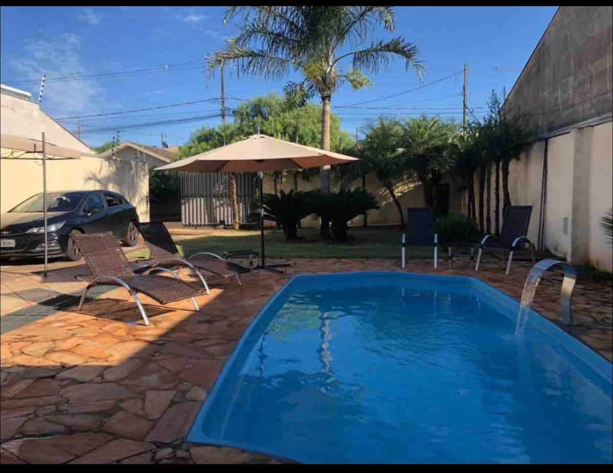 A clear pool is situated in a paved outdoor area, surrounded by lounge chairs and a shaded umbrella. Lush greenery and palm trees provide a serene backdrop, enhancing the space. The external environment is tranquil, featuring a vehicle parked nearby.