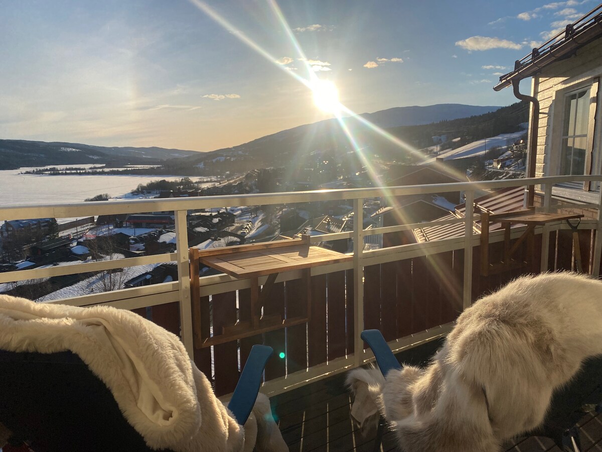 A spacious balcony features wooden furniture and cozy blankets, overlooking a serene landscape of snow-covered hills and valleys. The sun sets in the background, casting warm rays across the area, while distant trees and roofs are visible, creating a tranquil atmosphere.