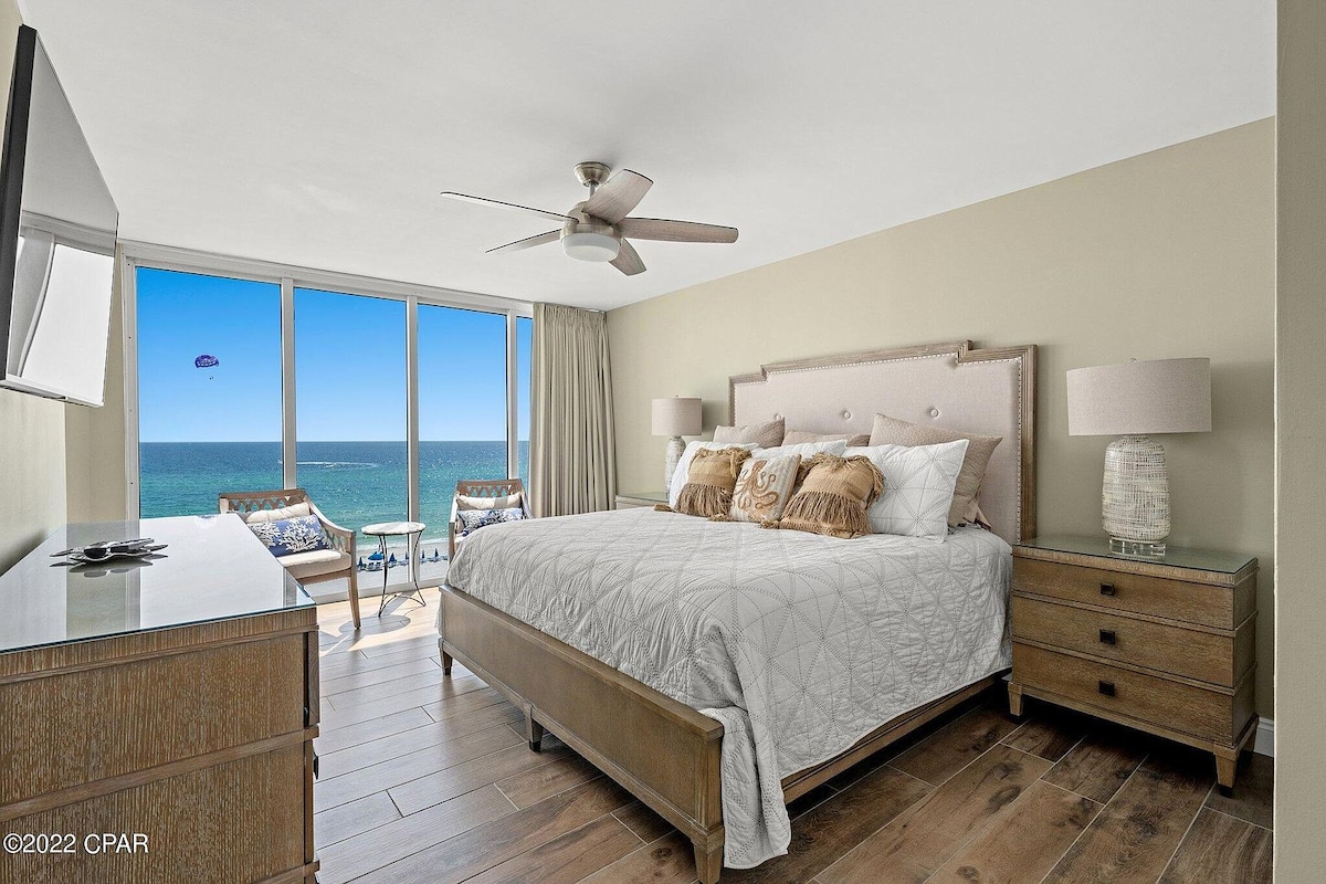 A bright bedroom is designed with large windows offering views of the Gulf of Mexico. A spacious bed is centered in the room, flanked by matching nightstands with lamps. Light wood flooring complements the serene atmosphere, and a TV is mounted on the wall opposite the bed.