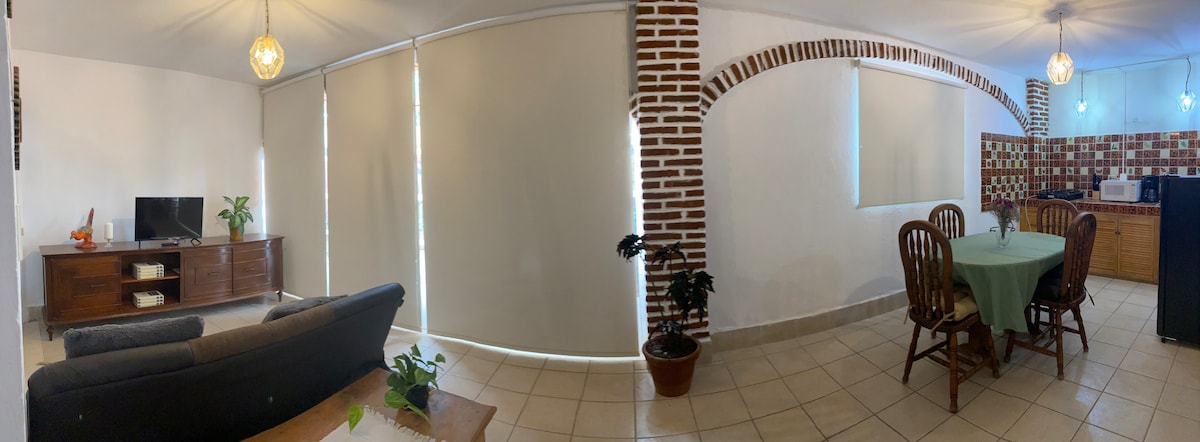 A spacious living area features a comfortable seating arrangement with a television unit. Natural light filters through large blinds, complementing the brick accents in the room. A dining space with a round table and chairs is visible, along with a kitchenette area showcasing wooden cabinetry.