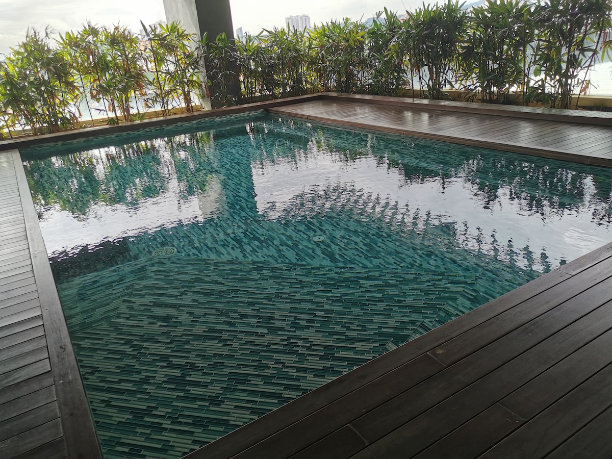 An outdoor swimming pool is set within a wooden deck area, surrounded by lush greenery. The pool features a patterned tile design and reflects the sky above, creating a serene atmosphere. Sunlight plays on the water's surface, enhancing the tranquil environment.