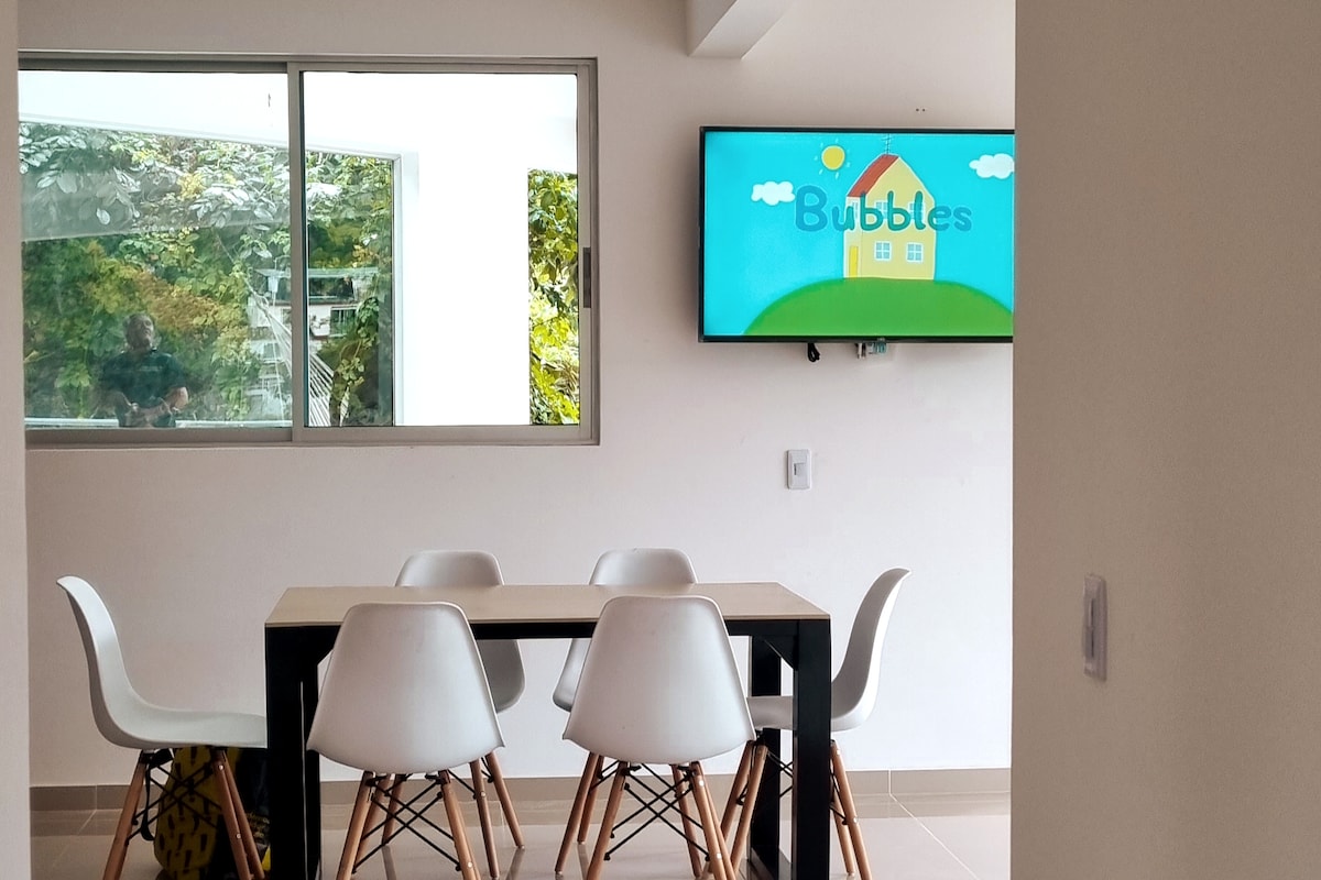 A dining area is presented with a light wooden table accompanied by six white chairs. A large window allows natural light to fill the space, while a television is mounted on the wall, displaying a colorful children's program.