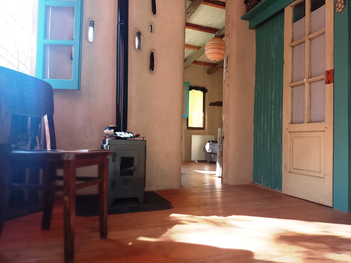 The interior space showcases a warm, wooden floor and walls with a rustic finish. A vintage chair is positioned beside a small stove. Doorways feature bright turquoise frames, and light filters through windows adorned with light-colored curtains, creating an inviting atmosphere throughout the home.
