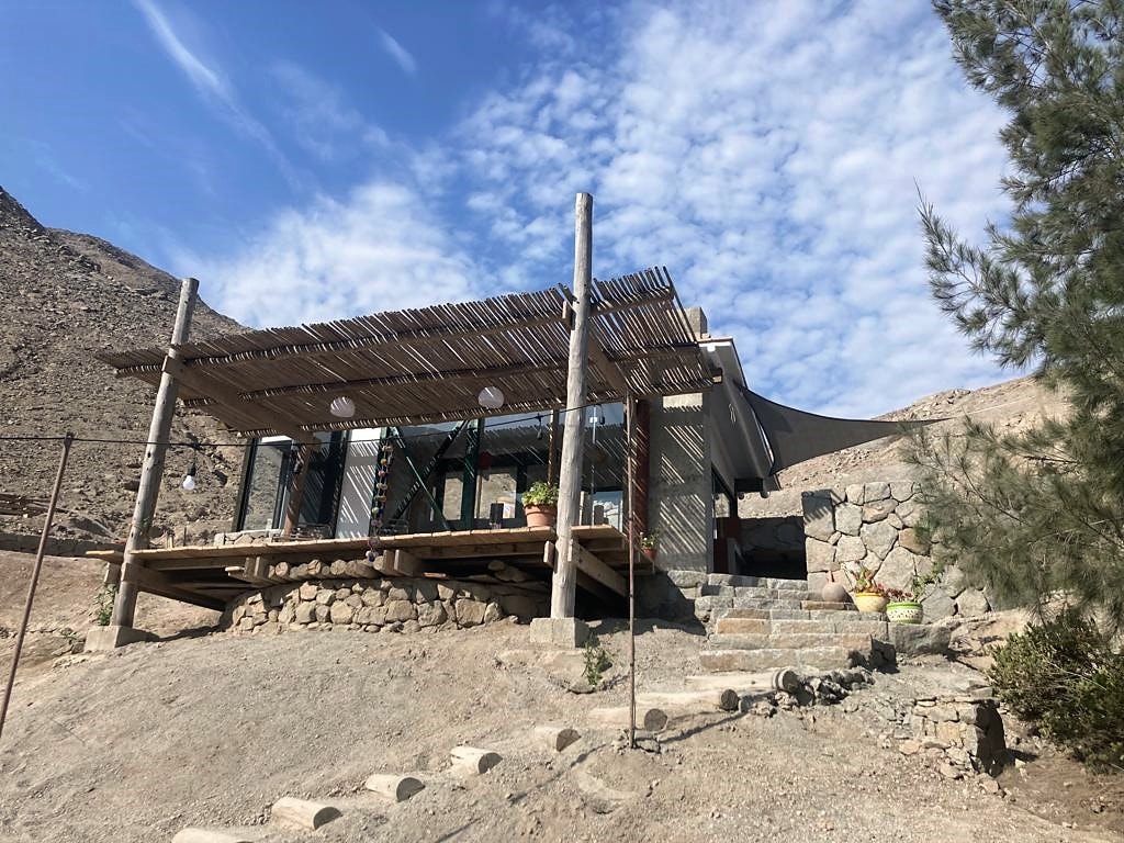 A cabin is situated on a hillside, featuring a wooden deck supported by sturdy posts. Large glass windows enhance the structure, providing unobstructed views of the surrounding landscape. Steps made of stone are lined along the pathway leading to the entrance.