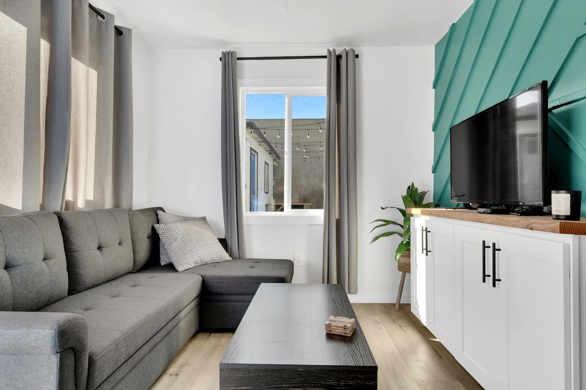 A modern living area is presented with a gray sectional sofa and a sleek coffee table. A television is mounted on a white cabinet against a teal wall. Natural light enters through a large window, draped with light gray curtains.