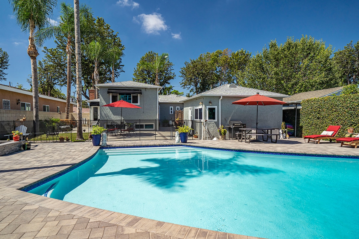 A large, clear saltwater pool is surrounded by a spacious patio and palm trees. The backyard features comfortable lounge chairs, umbrellas, and various potted plants. The home is visible in the background, showcasing a relaxed outdoor area perfect for families.