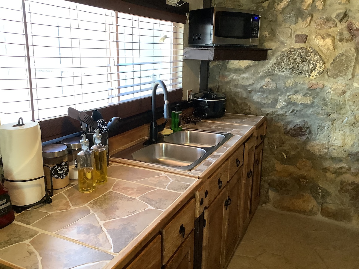 A kitchenette features a stone wall and a countertop with double sinks. Kitchen essentials, including bottles of oil and spices, are neatly arranged. A microwave is mounted above for convenience, and natural light filters through the window, highlighting the rustic ambiance.