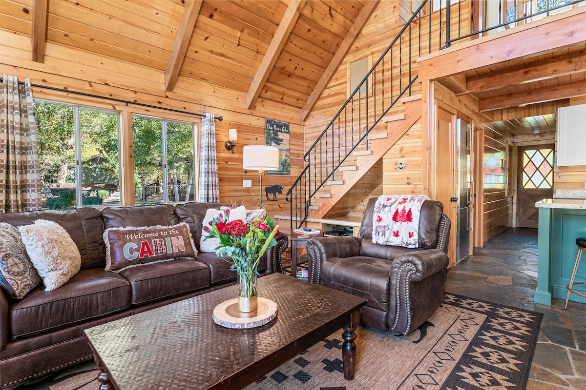 A cozy living area features a dark leather couch and reclining chair, surrounded by wooden walls with a warm tone. A coffee table holds a vase of flowers, while large windows allow natural light to illuminate the space. A staircase leads to the upper level.