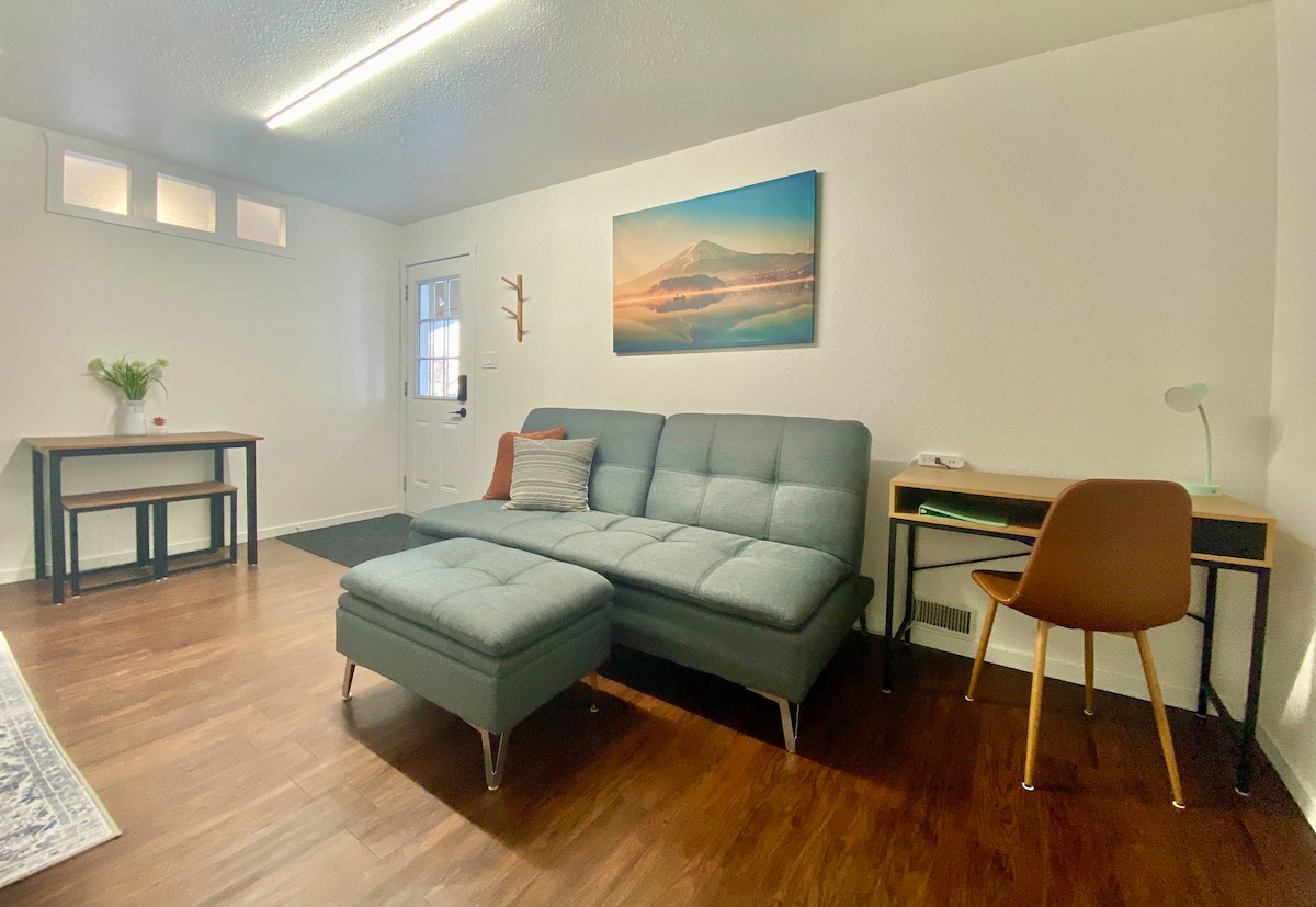 A compact living area features a modern sectional sofa and a small dining table. A desk with a chair provides a dedicated workspace, while a scenic wall art piece adds a touch of nature. Natural light enters through a door and nearby windows.