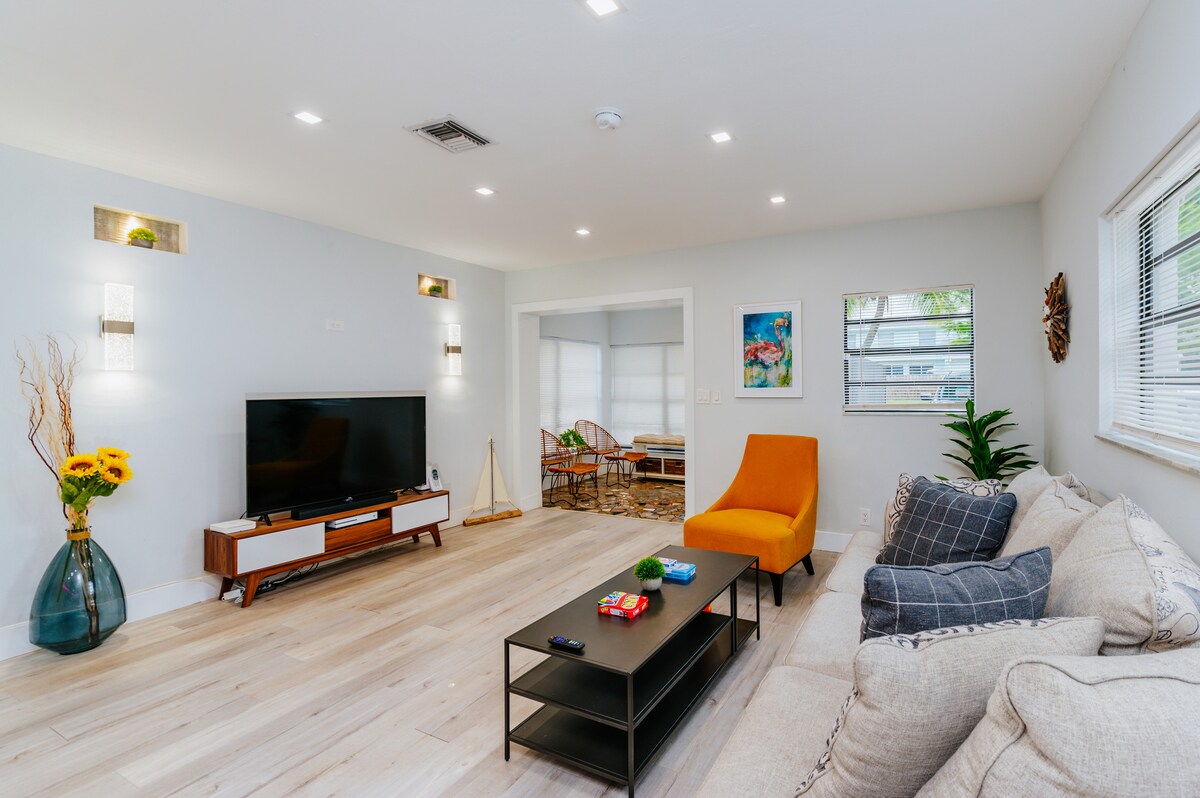 A bright and open living area features light-colored wooden flooring and soft grey walls. A comfortable sectional sofa is positioned invitingly, accompanied by an orange accent chair. A flat-screen TV rests on a contemporary media console, with decorative accents and plants adding subtle charm.