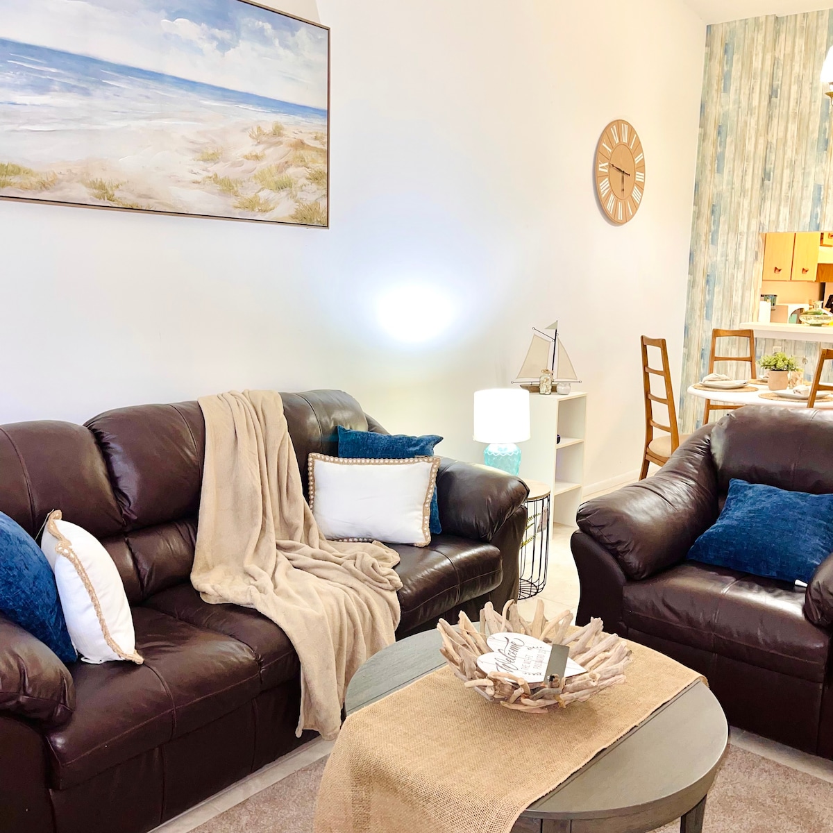 A comfortable living area is presented with dark leather sofas adorned with blue and white cushions. A textured tan blanket drapes over one sofa, and a wooden coffee table featuring a decorative centerpiece is positioned in front. Soft lighting and a beach-themed painting enhance the coastal vibe.
