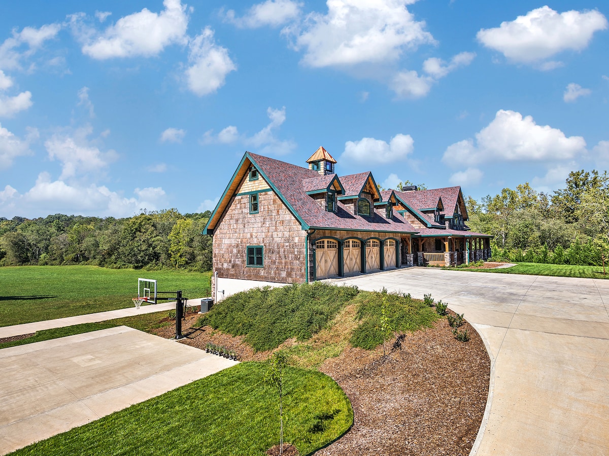 A large, rustic-style house is set against a backdrop of green fields and trees. The exterior features a combination of stone and wood materials, with multiple gabled roofs and decorative elements. A spacious driveway leads to the entrance, providing ample parking space.
