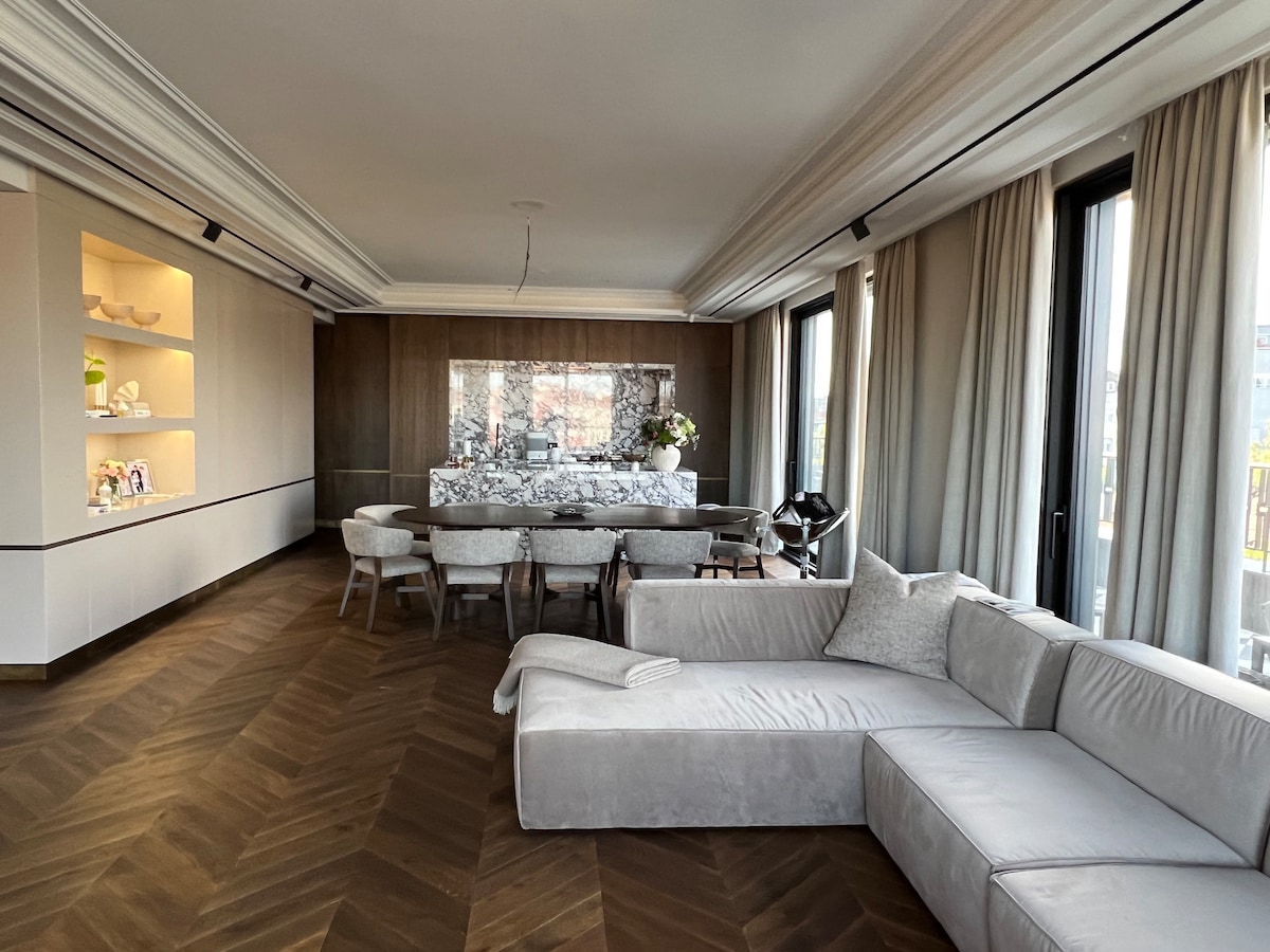 A spacious living area features a modern sectional sofa in a light color. In the background, a dining table surrounded by several chairs is visible, while natural light filters through large floor-to-ceiling windows adorned with soft curtains. Elegant design elements create a sophisticated ambiance.