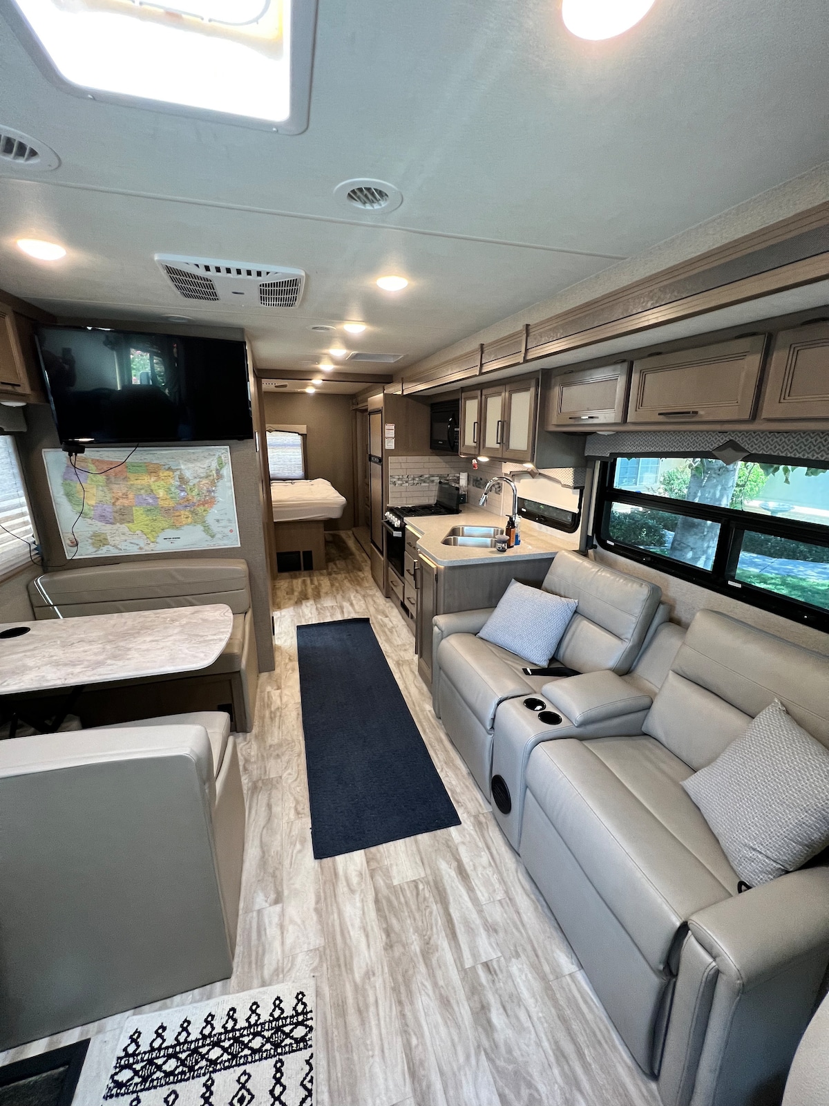 An interior view of a motorhome features a light-colored seating area with cup holders. A dining table is set nearby, alongside a well-equipped kitchen. The layout includes a spacious bed located towards the rear, with natural light filtering through the windows.