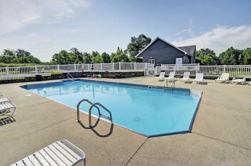 An outdoor swimming pool features clear blue water, surrounded by a spacious concrete deck. Several lounge chairs are positioned around the pool, with a white fence providing a boundary. Lush greenery is visible in the background, adding a sense of tranquility.