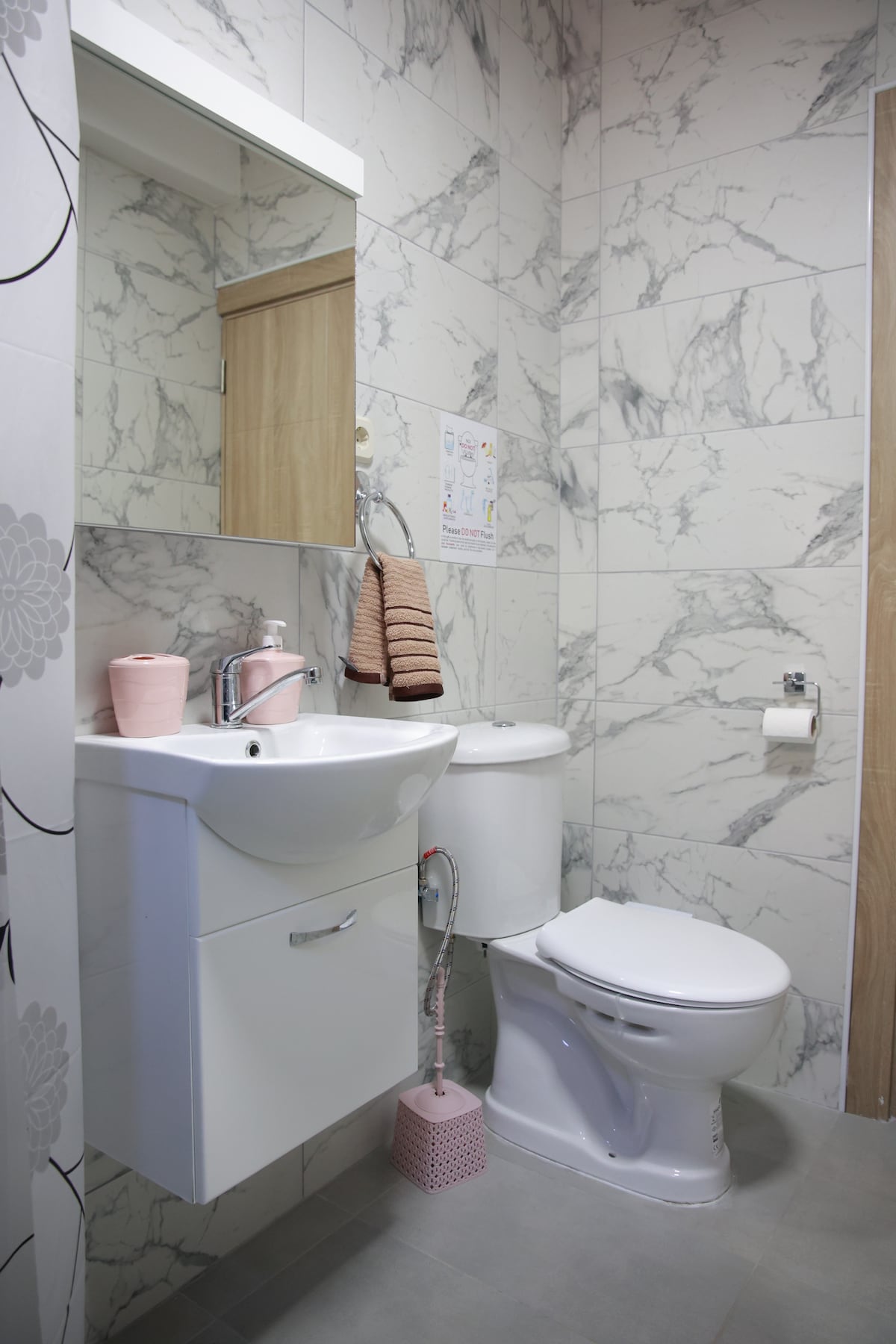 A modern bathroom features marble-patterned tiles in neutral tones. A sleek sink is accompanied by a mirror above, with toiletries and accessories arranged neatly. A compact toilet is positioned beside the sink, and a towel is hung on a rail for convenience.