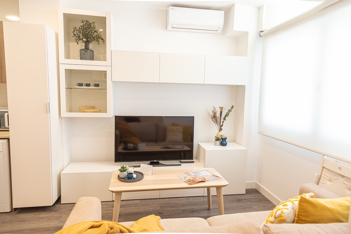 A modern living space features a minimalist design with a light-colored sofa adorned with a yellow throw. A sleek coffee table holds decorative items. A flat-screen television is mounted on a white wall unit, which includes shelves with plants and ceramics, enhancing the room's bright atmosphere.