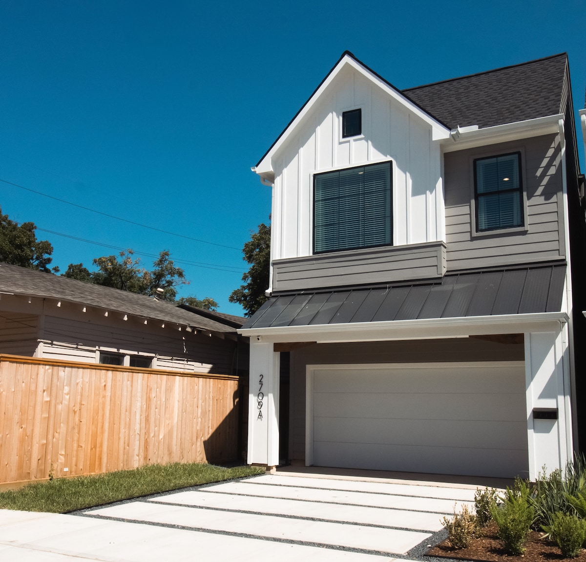 The exterior of a modern residential home features a clean facade with a combination of siding and large windows. A spacious driveway leads to a garage door, while a wooden fence provides privacy on one side. Natural landscaping complements the architecture under a clear blue sky.
