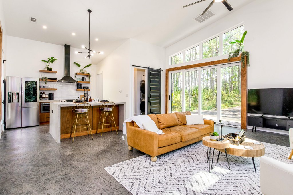 A modern living area features a large brown sofa situated on a patterned rug. A spacious kitchen is equipped with stainless steel appliances and bar seating. Large windows allow natural light to illuminate the space, offering views of the surrounding greenery.