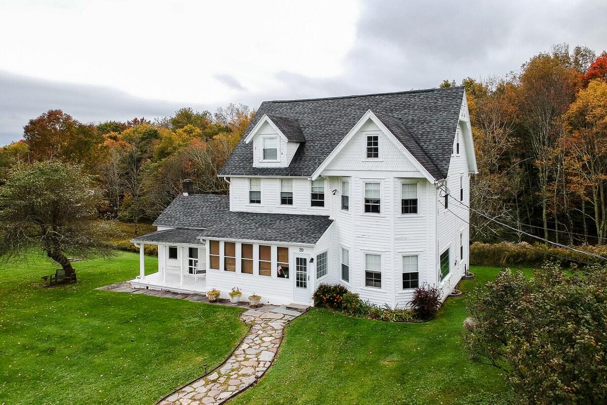 The large three-story farmhouse is enveloped by green lawns and trees, showcasing a crisp white exterior. A stone pathway leads to the entrance, while large windows illuminate the front porch, integrated with the serene landscape of colorful foliage in the background.