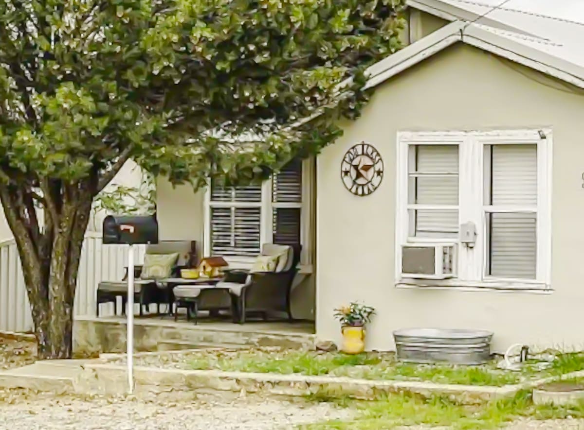The exterior of the casita features a welcoming porch with comfortable seating arrangements and decorative pillows. A clock is mounted on the wall beside the entrance. A shaded tree provides greenery, while a modern mailbox and a small planter add charm to the entrance.