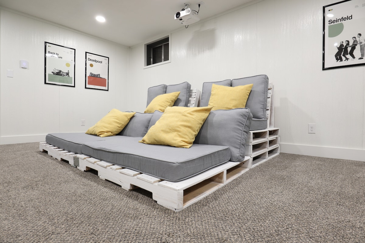 A casual seating area is created with a custom pallet couch, adorned with several yellow and gray throw pillows. A wall-mounted projector hangs above, ready for movie nights. The carpeted floor adds comfort to the inviting space.