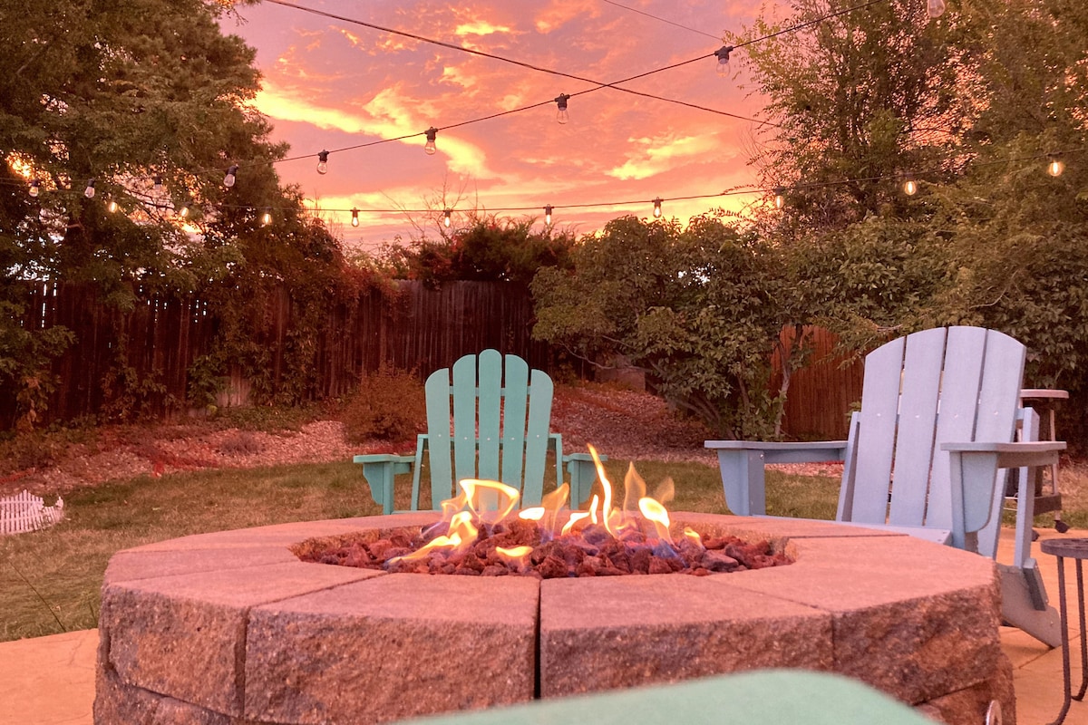 A gas fire pit sits at the center of a spacious backyard, surrounded by Adirondack chairs. The vibrant sunset casts warm hues across the sky, while string lights create an inviting atmosphere. Lush greenery and a wooden fence frame the scene, enhancing the sense of privacy.
