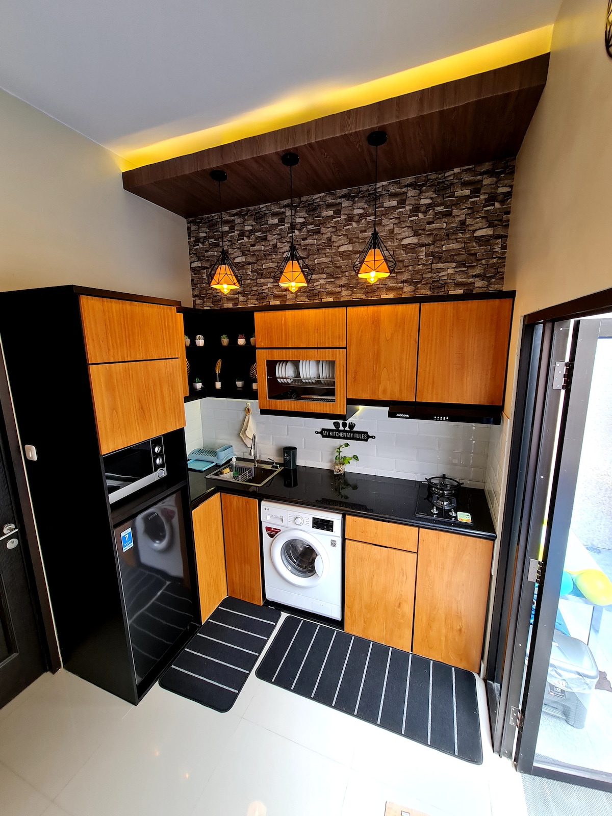 The kitchen features a modern design with wooden cabinetry and a dark countertop. A washing machine is integrated, alongside appliances including a refrigerator and stove. Decorative pendant lights hang from the ceiling, and a plant adds a touch of greenery to the space.