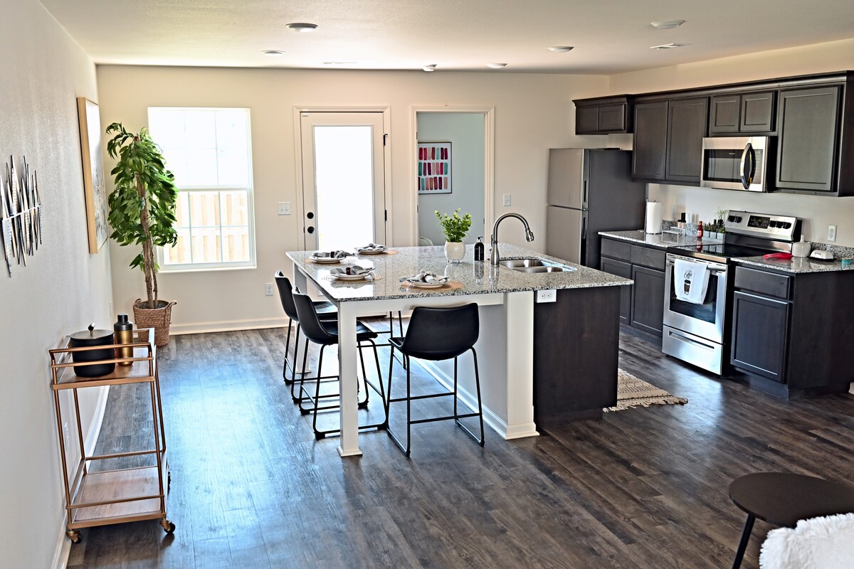 A modern kitchen and dining area features a large granite island with seating for four. Stainless steel appliances are showcased alongside stylish ebony cabinets. Natural light enters through the window, illuminating the space. A potted plant adds a touch of greenery to the room.