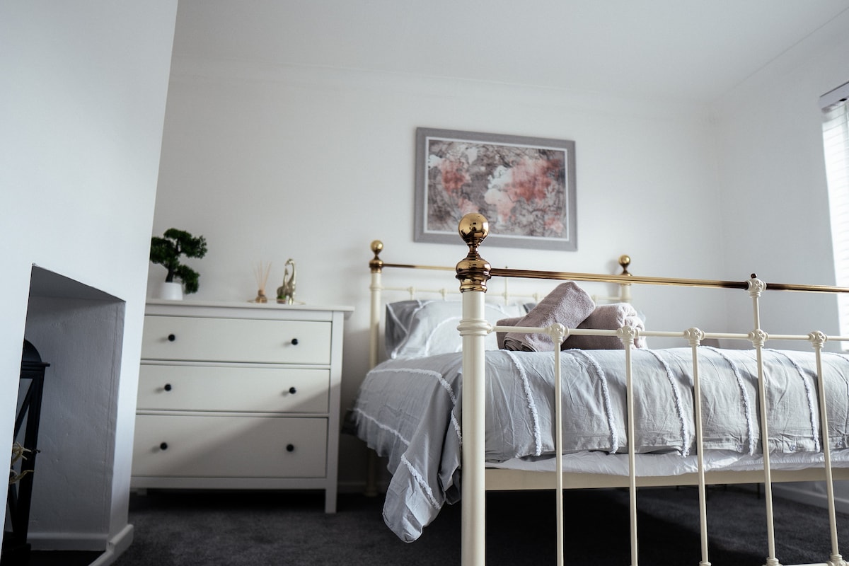A spacious bedroom is featured, showcasing a metal-framed bed with a soft, neutral-toned bedding. A white dresser is positioned nearby, accompanied by a simple decorative element. Natural light enters through a window, illuminating the subtle color palette of the room.