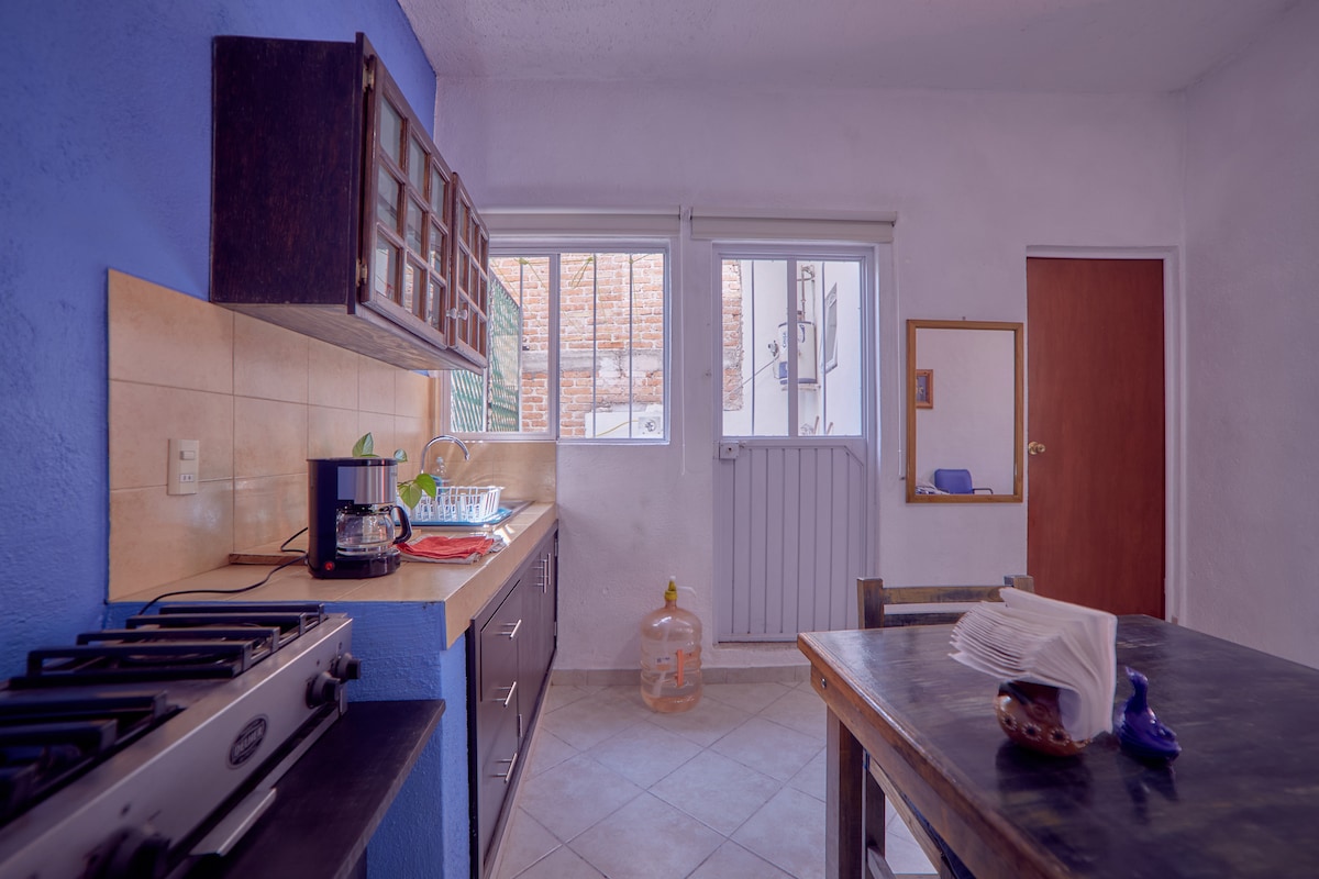 A well-organized kitchen is presented with a blue accent wall and wooden cabinetry. Natural light floods through window units, illuminating the counter space which includes a stove and kitchen appliances. A wooden dining table is visible, accompanied by neutral-colored tableware.