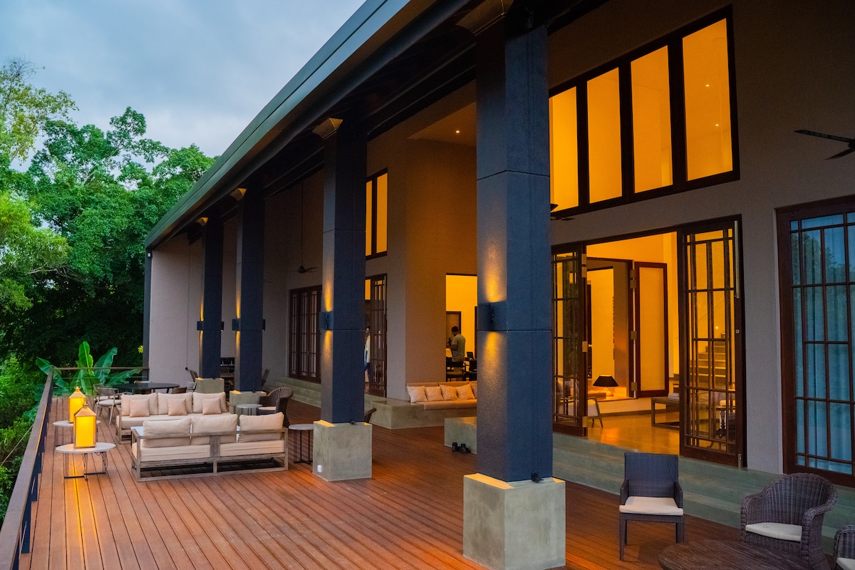 A spacious deck extends from the villa, featuring comfortable seating arrangements and elegant lighting. Large glass doors lead into the interior, framed by tall windows that allow natural light to fill the space. Lush greenery surrounds the area, enhancing the serene setting.