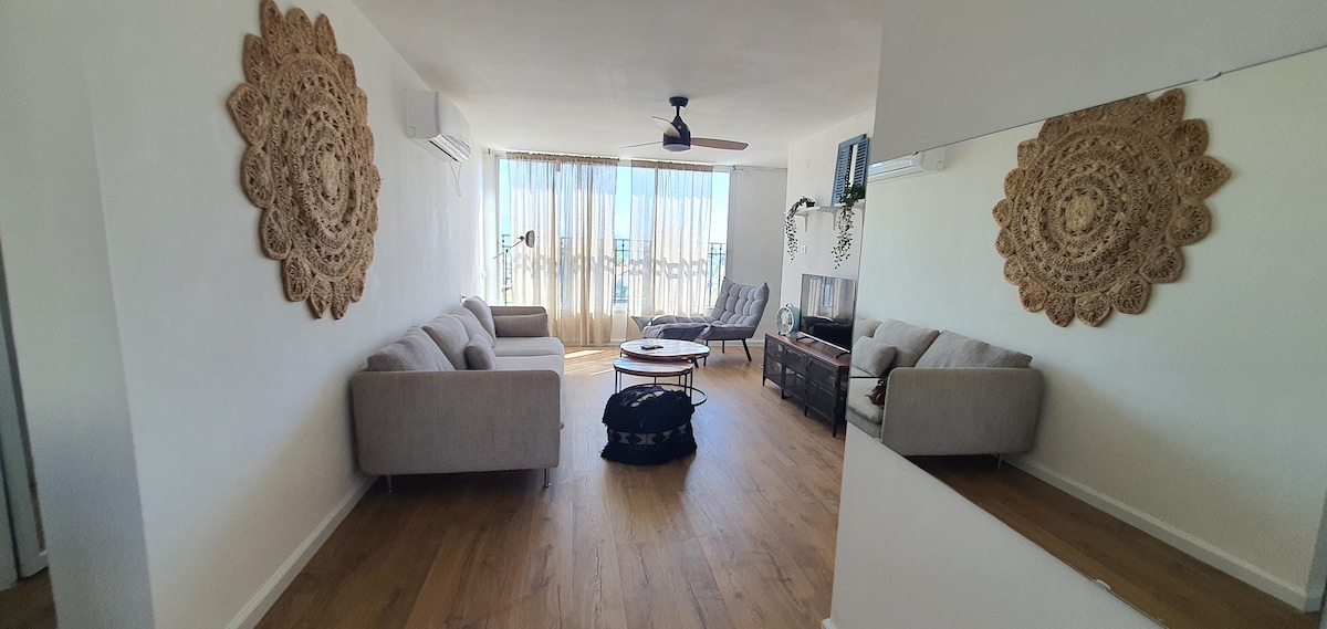 A spacious living area features light-colored sofas surrounding a round coffee table. Decorative wall hangings add texture, while a large window offers bright natural light and views. A wall mirror reflects the open space, contributing to a feeling of airiness.