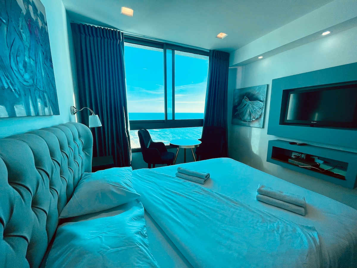 A well-lit bedroom features a spacious bed with neatly arranged towels. Large windows offer a clear view of the exterior, allowing natural light to fill the room. A seating area with a small table and an armchair is positioned near the window, complemented by tasteful artwork on the walls.