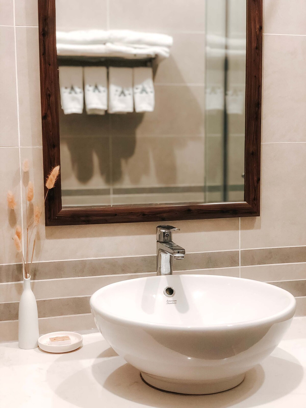 A modern bathroom features a round white sink positioned below a wooden-framed mirror. Soft neutral tiles create a calming backdrop, while a small vase with dried plants adds a decorative touch. Several neatly arranged towels hang in the background, complementing the serene atmosphere.