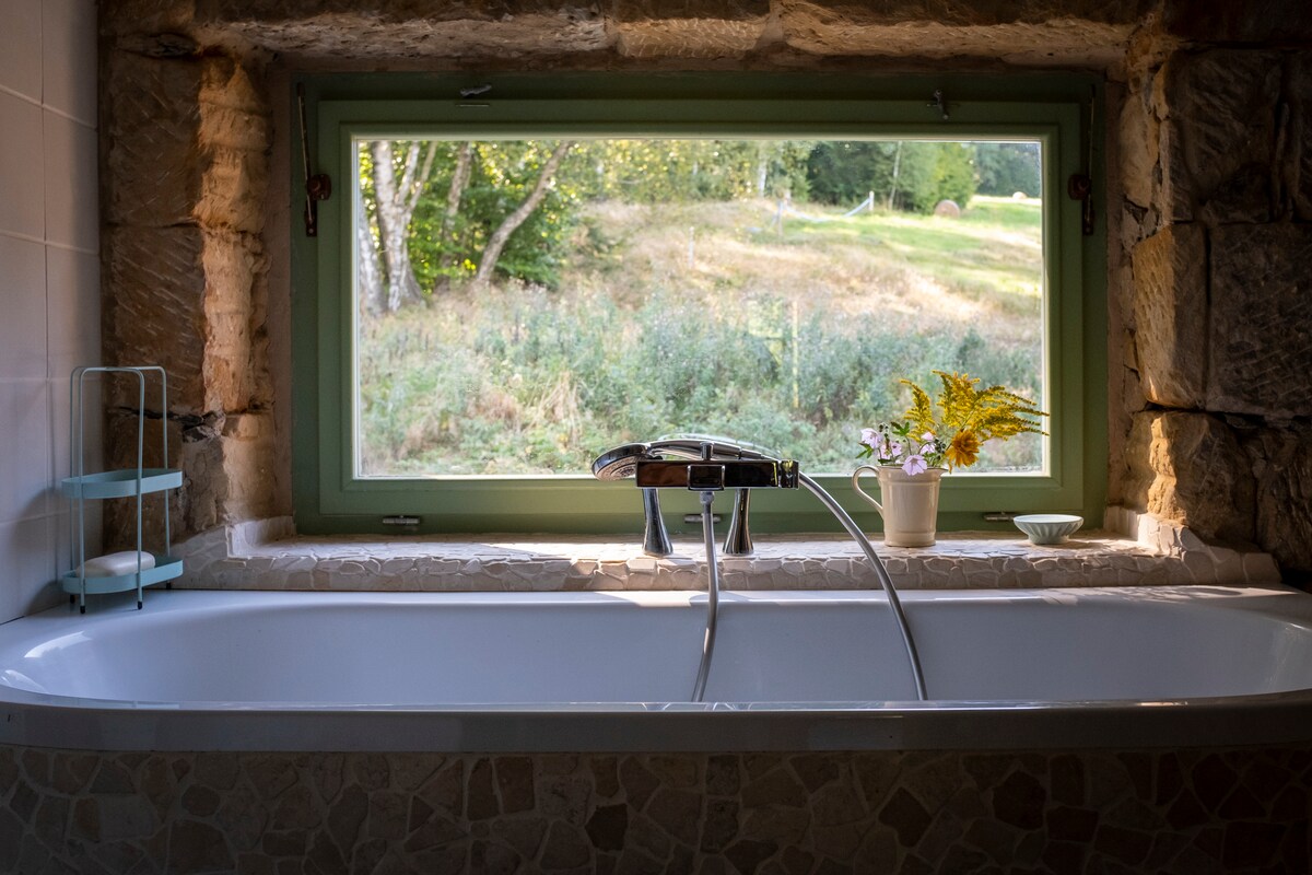 A modern bath is positioned beneath a large window framed by stone, offering views of the lush outdoor scenery. A small shelf holds a decorative potted plant and dish, enhancing the serene atmosphere.