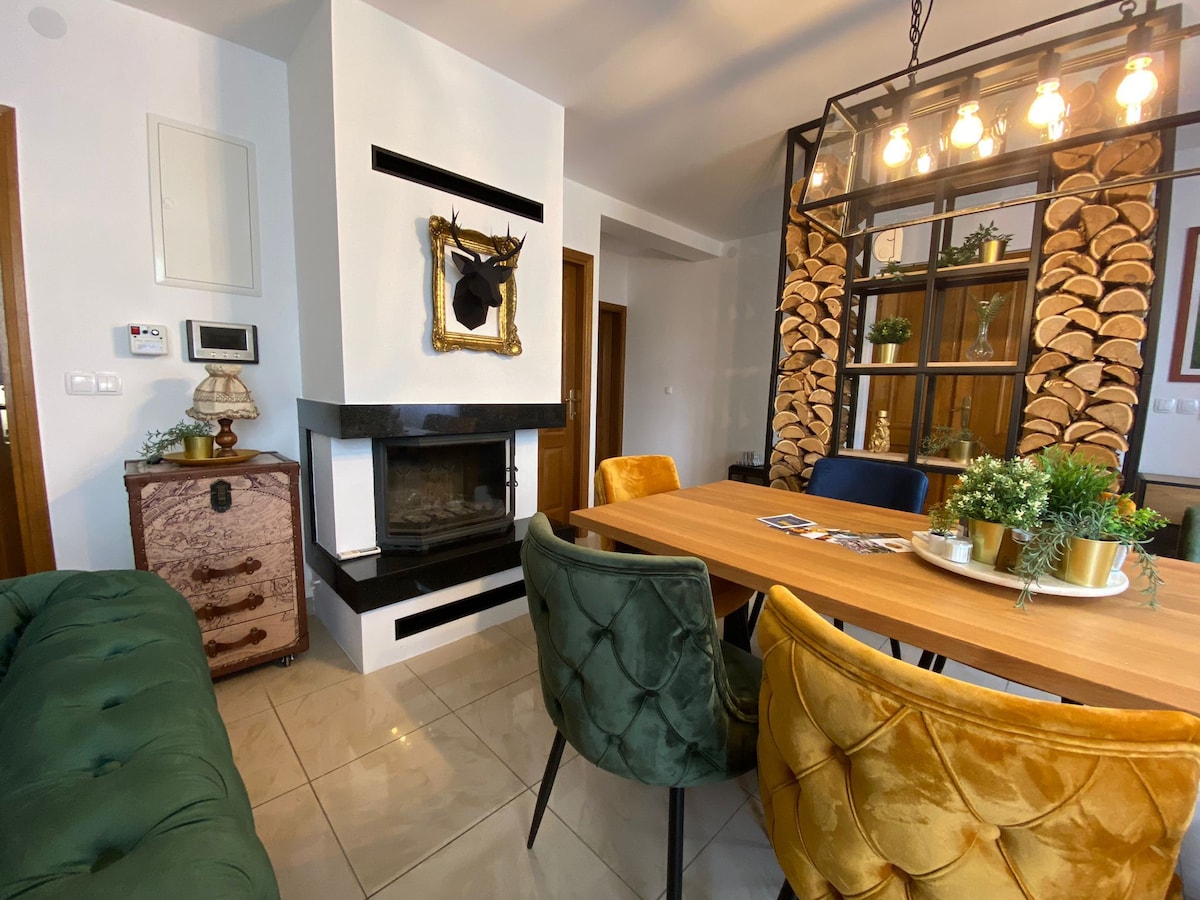 A modern dining area features a wooden table surrounded by plush, upholstered chairs in varied colors. A stylish glass and metal light fixture hangs above, while a cozy fireplace is set into the wall, adorned with decorative logs and an ornate mirror.