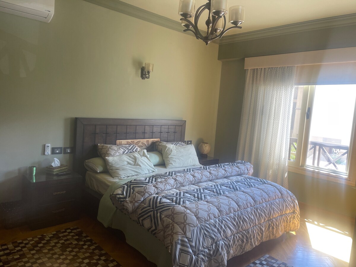 A well-lit bedroom features a spacious bed with a patterned duvet and decorative pillows, positioned near a large window. Soft green walls and a stylish chandelier enhance the room's decor, while a textured rug adds warmth to the hardwood floor.