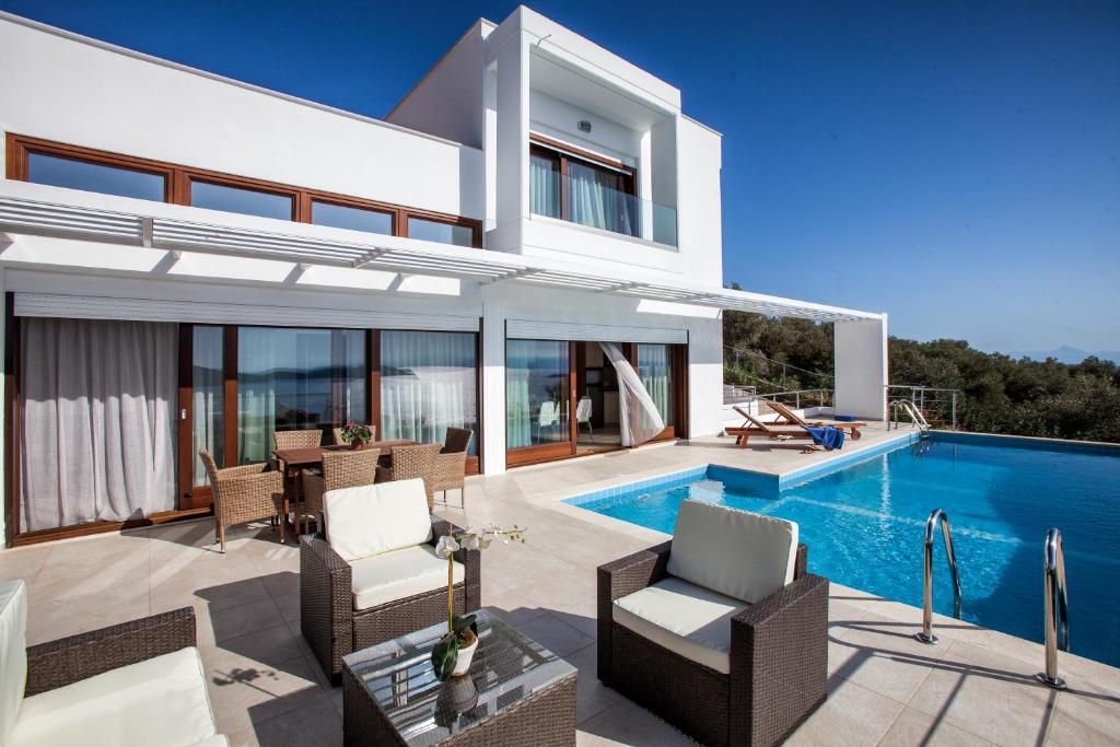 An outdoor seating area features light-colored furniture arranged around a glass table, set beside a swimming pool. Large glass doors provide access to the villa's interior. A canopy extends over the space, offering shade against the clear blue sky.