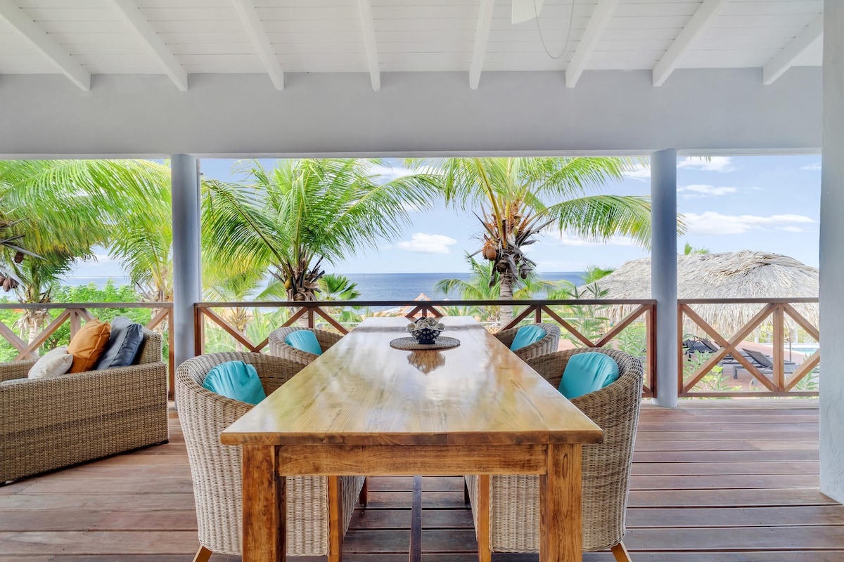 A spacious outdoor dining area features a wooden table surrounded by four wicker chairs, accented with light blue cushions. Lush palm trees and an ocean view are visible in the background, creating a serene setting for meals and relaxation.