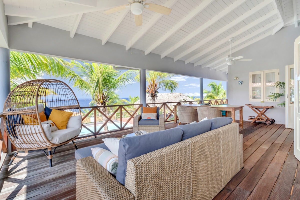 A spacious rear deck is lined with natural wood, offering views of palm trees and the ocean. A comfortable sofa is complemented by an inviting egg chair adorned with bright cushions. A dining table is positioned nearby, perfect for enjoying meals in the open air.