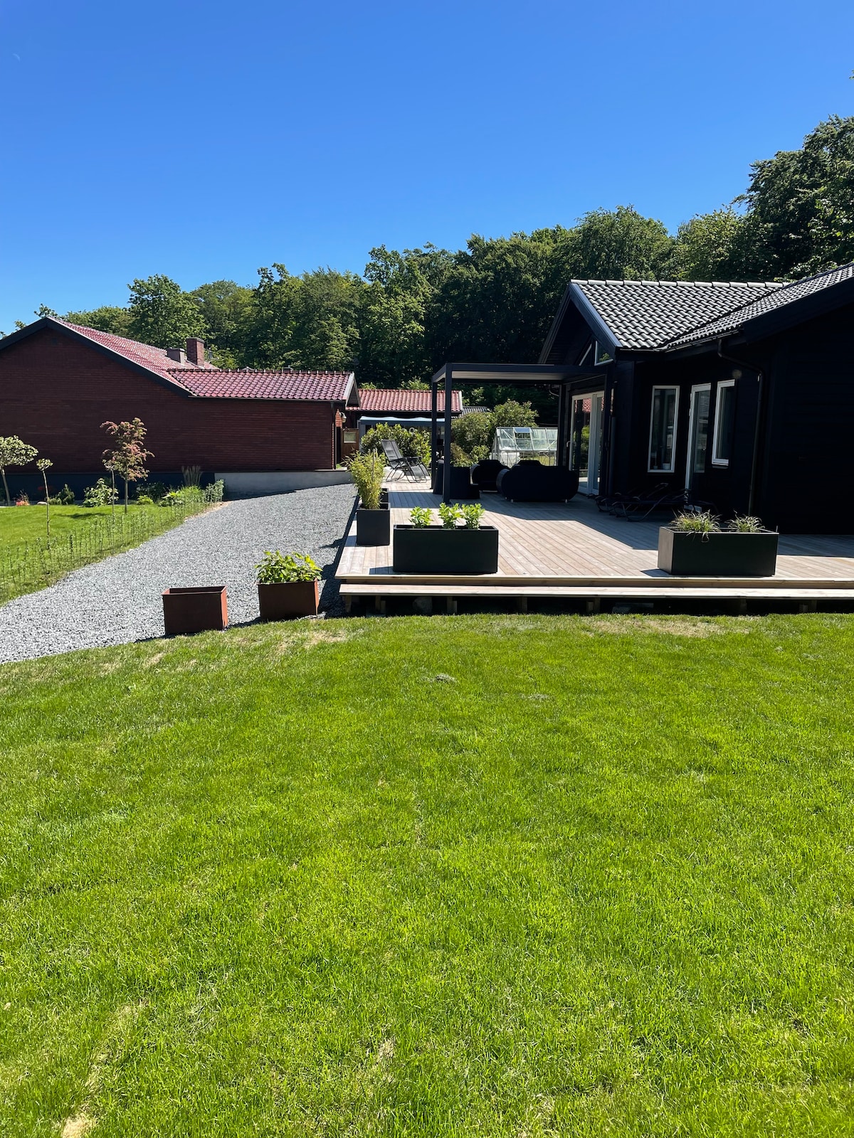 A well-maintained exterior features a sunny lawn bordered by gravel pathways and planters. The deck is adorned with green plants, while the nearby houses are nestled among trees, contributing to a peaceful and natural setting.