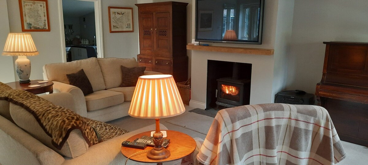 A cozy sitting room is presented with soft beige sofas, a wooden coffee table, and a warm log burner. A television is mounted above a simple mantel, and ambient light is provided by lamps with striped shades, creating a comfortable space for relaxation.